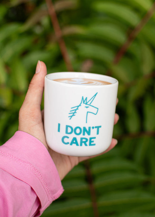 In der Hand wird eine Tasse von J.Clay „I don’t Care“ gehalten; der Schriftzug „I DON’T CARE“ ist zu sehen, im Hintergrund ist Grün zu erkennen.