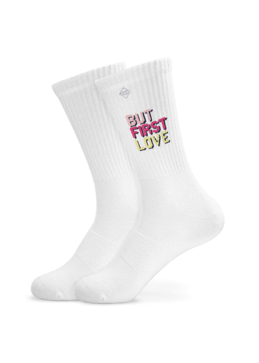 Ein Paar weiße J.Clay Tennissocken mit gerippten Bündchen, auf denen der Satz "BUT FIRST LOVE" in fetten, bunten Buchstaben oben aufgestickt ist.