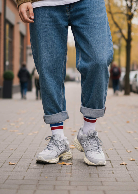 Eine Person mit Jeanshose und New Balance Schuhen trägt die weißen Tennissocken mit rot blauen Streifen