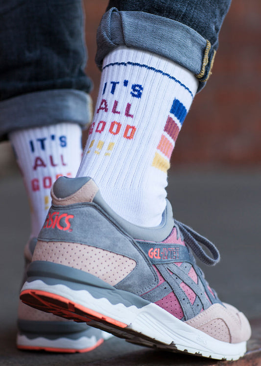 Nahaufnahme einer Person in hochgekrempelten Jeans, grauen und rosafarbenen Asics-Turnschuhen und weißen Tennissocken von J.Clay mit buntem Text und Balken, auf denen in blauen, roten und gelben Buchstaben ITS ALL GOOD steht.
