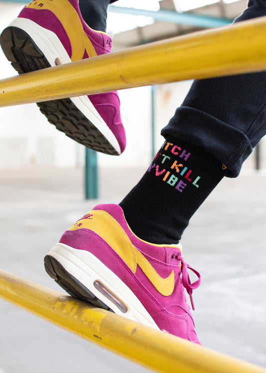 Nahaufnahme einer Person in schwarzen J.Clay-Tennissocken mit buntem "WITCH DONT KILL MY VIBE"-Schriftzug, magentafarbenen und gelben Nike-Turnschuhen, die auf einem gelben Metallgeländer in einer Halle steht.