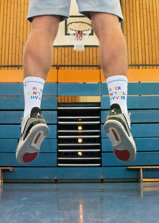 Eine Person springt in einer Turnhalle und trägt weiße J.Clay-Tennissocken mit der Aufschrift "Bitch Don't Kill My Vibe", graue Shorts und schwarz-weiße Turnschuhe. Im Hintergrund sind ein Basketballkorb und eine blaue Tribüne zu sehen.