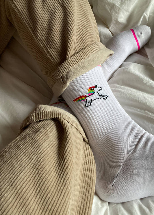 Jemand, der eine beigefarbene Cordhose und weiße J.Clay-Tennissocken mit einem kleinen Regenbogen-Einhornmuster trägt, sitzt auf einem Bett mit weißen Laken.
