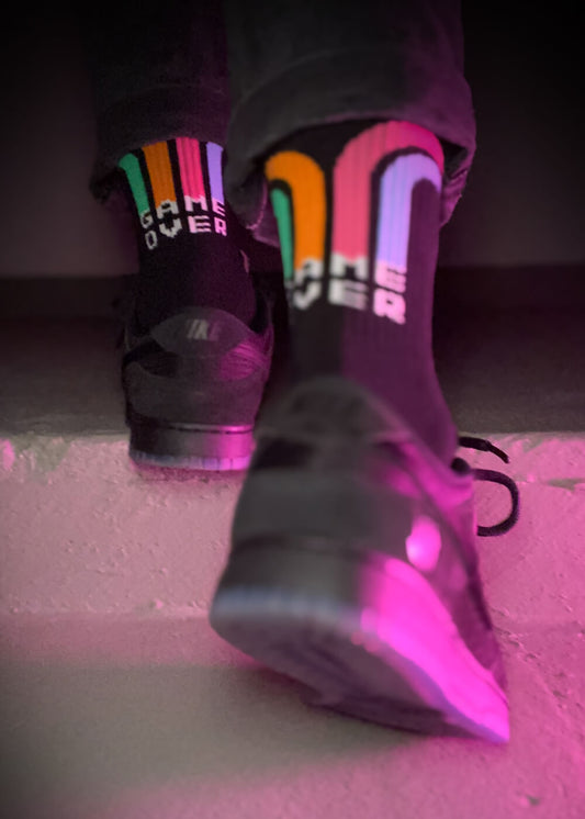 Eine Person in schwarzen Turnschuhen und J.Clay Tennissocken - schwarze Socken mit bunten Streifen und der Aufschrift "GAME OVER" - geht eine Treppe hinauf, die von rosa und violettem Licht beleuchtet wird.