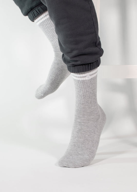 Eine Person trägt eine dunkelgraue Jogginghose von J.Clay und hellgraue Tennissocken mit weißen Streifen. Sie sitzt auf einer weißen Fläche, hat die Füße leicht gekreuzt und nur die Unterschenkel sind sichtbar.