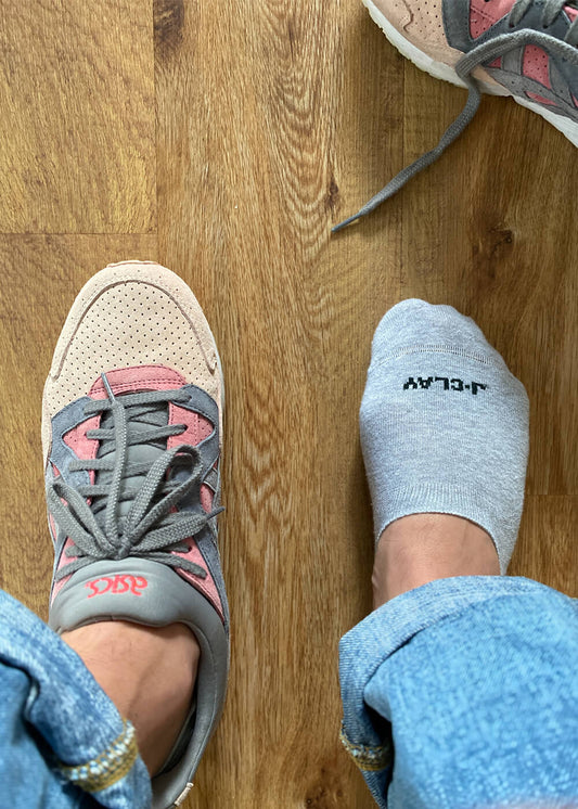Eine Person in blauer Jeans hat einen Fuß in einem rosa-grauen ASICS-Sneaker, während der andere eine graue J.Clay-Socke trägt - perfekt als unsichtbare Füßlinge, die nicht rutschen. Ein weiterer Turnschuh ist teilweise auf dem Holzboden zu sehen.