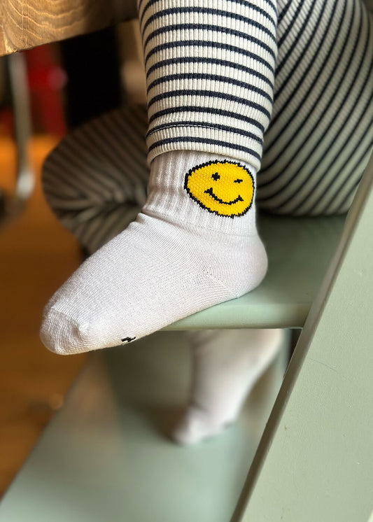 Ein Kind, das J.Clay Baby Socken mit einem gelben Smiley-Gesicht und schwarz-weiß gestreiften Leggings trägt, sitzt auf einem hellgrünen Stuhl. Ideal für Kinder Socken 0-4 Jahre, diese Socken bringen spielerischen Charme zu jedem Look.