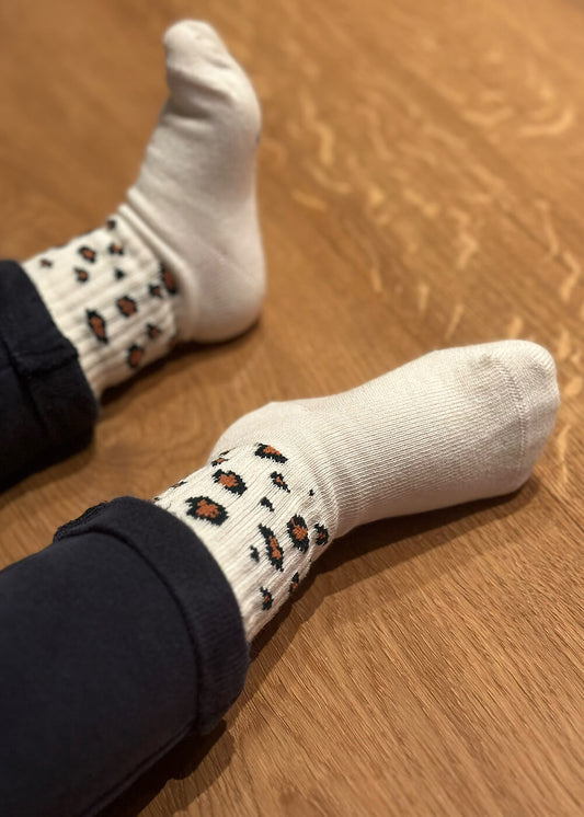 Ein Kind in dunkelblauer Hose liegt auf einem Holzboden und trägt J.Clay Baby Socken in Weiß mit schwarzem und braunem Leopardenmuster. Diese stylischen Kinder Socken sind für Kinder von 0-4 Jahren geeignet.