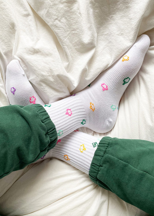 Eine Person in grünen Hosen sitzt im Schneidersitz auf einem weißen Bett und trägt weiße Tennissocken von J.Clay, die mit bunten Blumenmustern verziert sind.