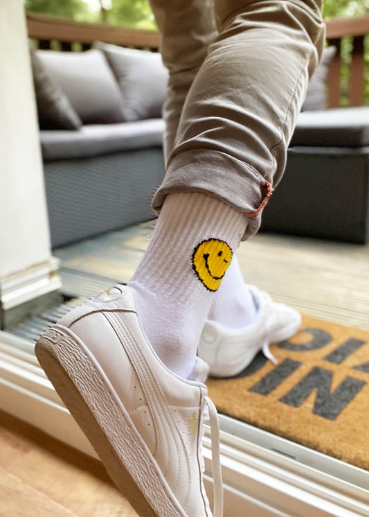 Eine Person in weißen J.Clay-Tennissocken mit einem gelben Smiley-Motiv und weißen Turnschuhen tritt auf eine Fußmatte, bekleidet mit hellbeigen Hosen, im Hintergrund sind Gartenmöbel zu sehen.