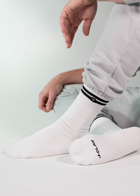 Eine Person in hellgrauer Hose sitzt mit einem angewinkelten Knie und trägt weiße J.Clay-Tennissocken mit schwarzen Streifen, einem kleinen Logo und "JCLAY" an der Sohle. Der Hintergrund ist einfarbig und hell gefärbt.
