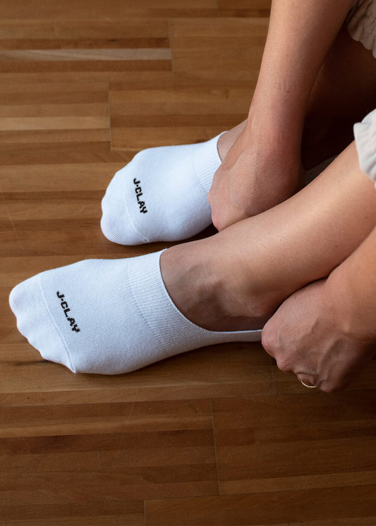 Eine Person sitzt auf einem Holzboden und trägt J.Clay unsichtbare Füßlinge mit dem Schriftzug "J.CLAY" in der Nähe der Zehen, wobei sie eine Socke -perfekt als rutschfeste Füßlinge- anpasst.