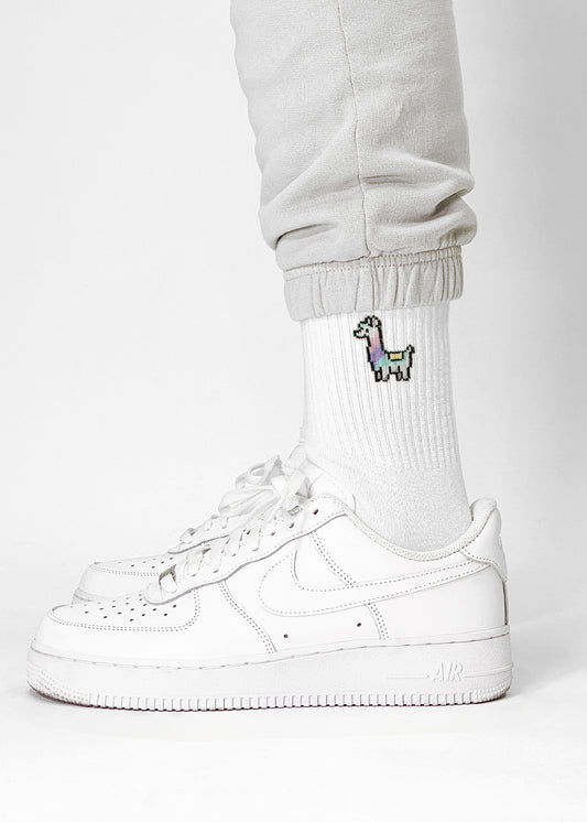 Eine Person trägt weiße Turnschuhe, helle Jogginghosen und J.Clay-Tennissocken mit einem kleinen verpixelten Lama-Muster in der Nähe des Knöchels vor einem schlichten weißen Hintergrund.